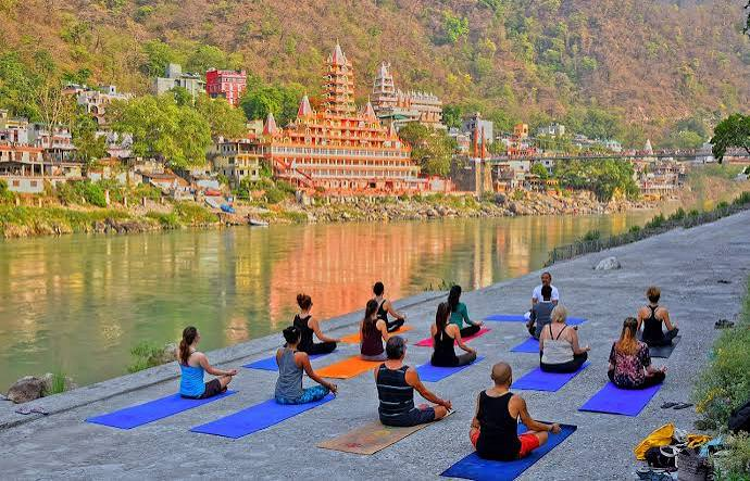 "Best Places to Visit in Uttarakhand – Tourists practicing yoga on the banks of the Ganges River in Rishikesh with temples and scenic hills in the background."